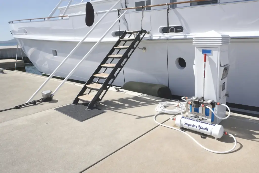 Compact watermaker unit and pressure vessel on a marina pontoon beside a yacht, hoses run to the dock pedestal.