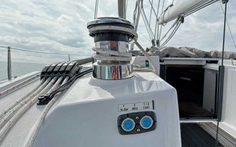 Polished electric sheet winch with white rope wraps on a modern sailing yacht's cockpit coaming, with the dual-speed control pad and its two backlit buttons below, rope clutches and side deck beyond.