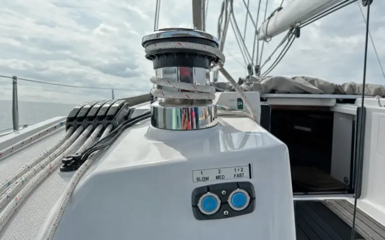 Polished electric sheet winch with white rope wraps on a modern sailing yacht's cockpit coaming, with the dual-speed control pad and its two backlit buttons below, rope clutches and side deck beyond.