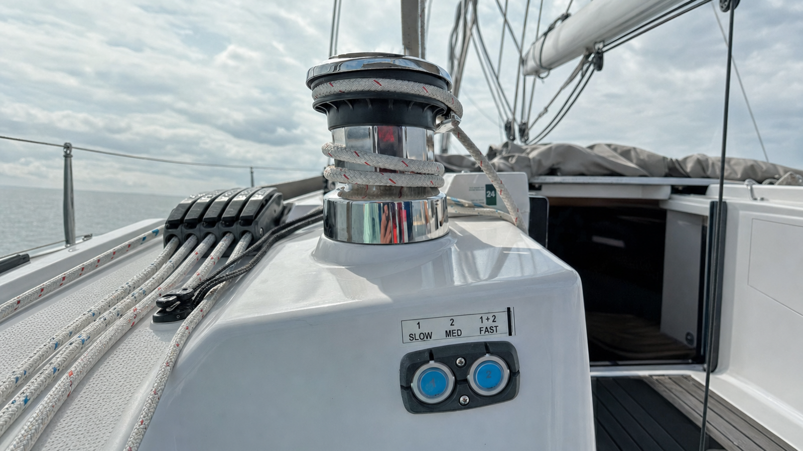 Polished electric sheet winch with white rope wraps on a modern sailing yacht's cockpit coaming, with the dual-speed control pad and its two backlit buttons below, rope clutches and side deck beyond.