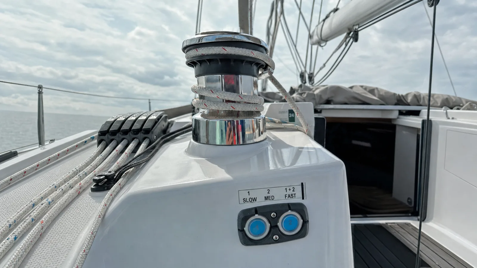 Polished electric sheet winch with white rope wraps on a modern sailing yacht's cockpit coaming, with the dual-speed control pad and its two backlit buttons below, rope clutches and side deck beyond.